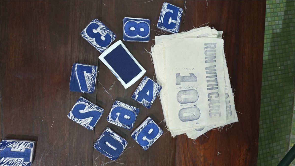 Hand-carved rubber-stamp letters and numbers spelling out RUN WITH CARE, an ink pad, and a stack of stamped fabric BIBs on a wooden table