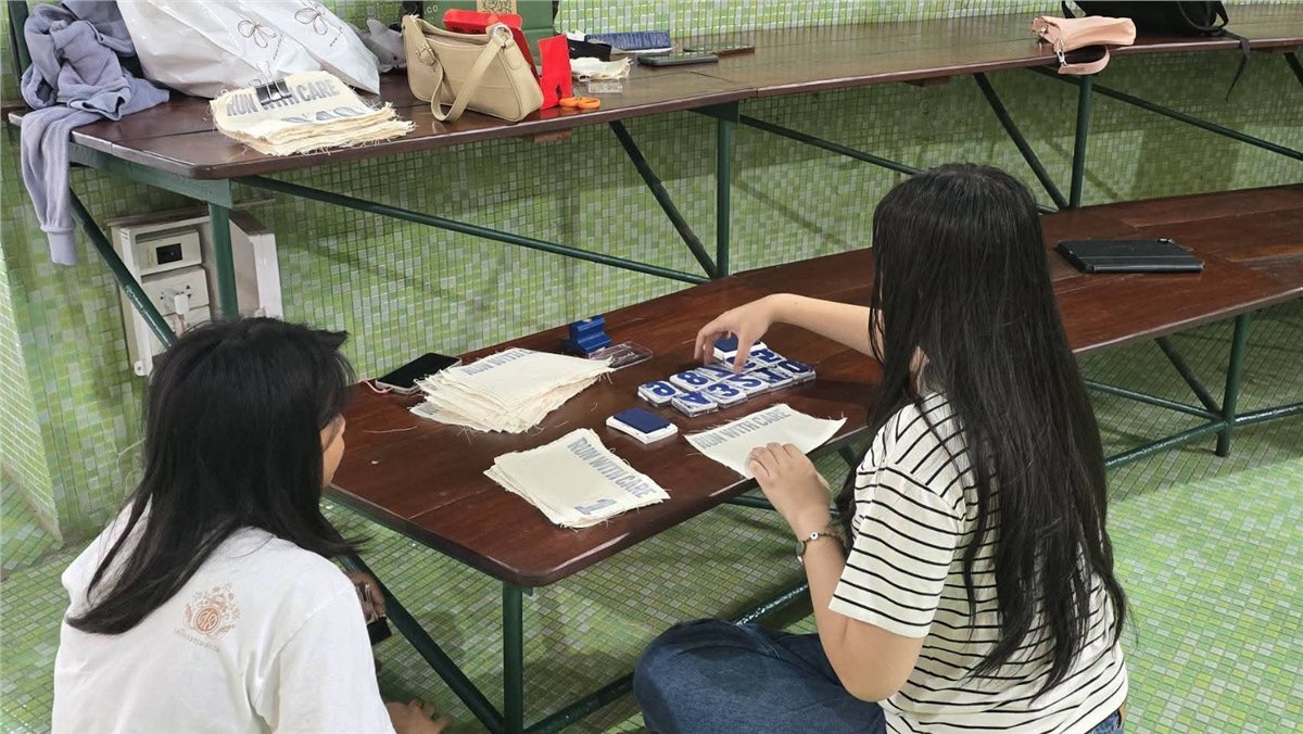 Two volunteers hand-stamping RUN WITH CARE BIB numbers onto fabric at a table
