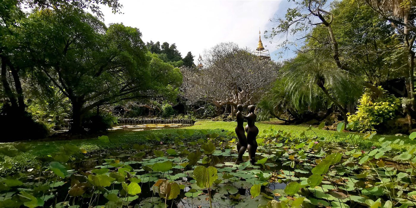 The gardens of Sathira-Dhammasathan — a lily-pad pond with a sculpture of two figures, frangipani and palm trees, and a golden chedi rising above the treetops